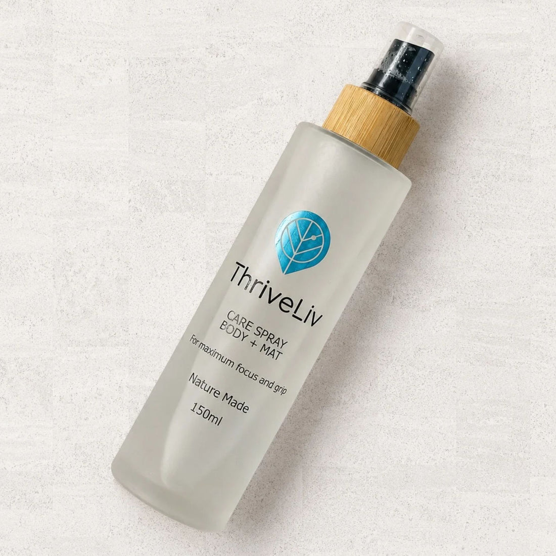 A 150ml bottle of ThriveLiv Care Spray Body + Mat on a textured light gray background. The bottle is frosted with a light wood-toned cap and a clear spray nozzle.