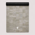 A rectangular yoga mat with a light gray stone tile pattern, partially unrolled to show its black textured underside, featuring the ThriveLiv logo at the bottom.